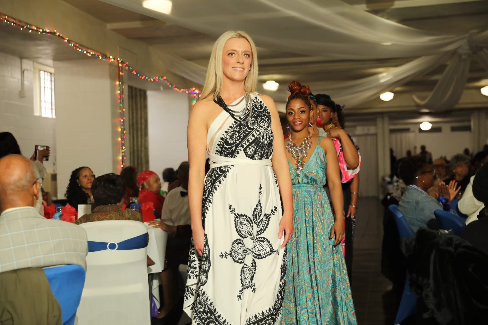 Model walking down a runway in a white patterned dress at a fashion event with an audience on either side.